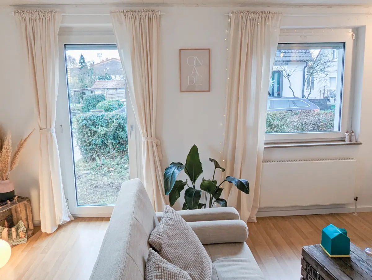 Helles Wohnzimmer mit großen Fenstern, Sofa und Pflanzen in gemütlicher Atmosphäre.