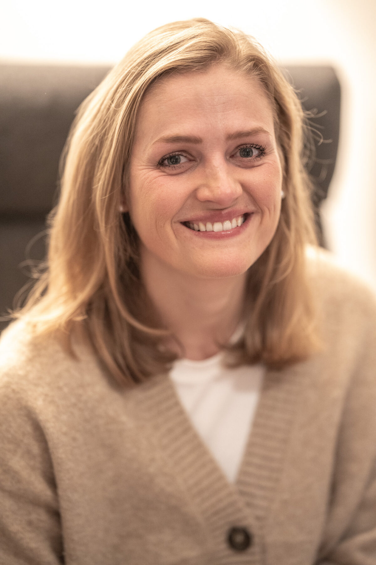 Eine lächelnde Frau in einem beige Pullover, sitzend im Büro.